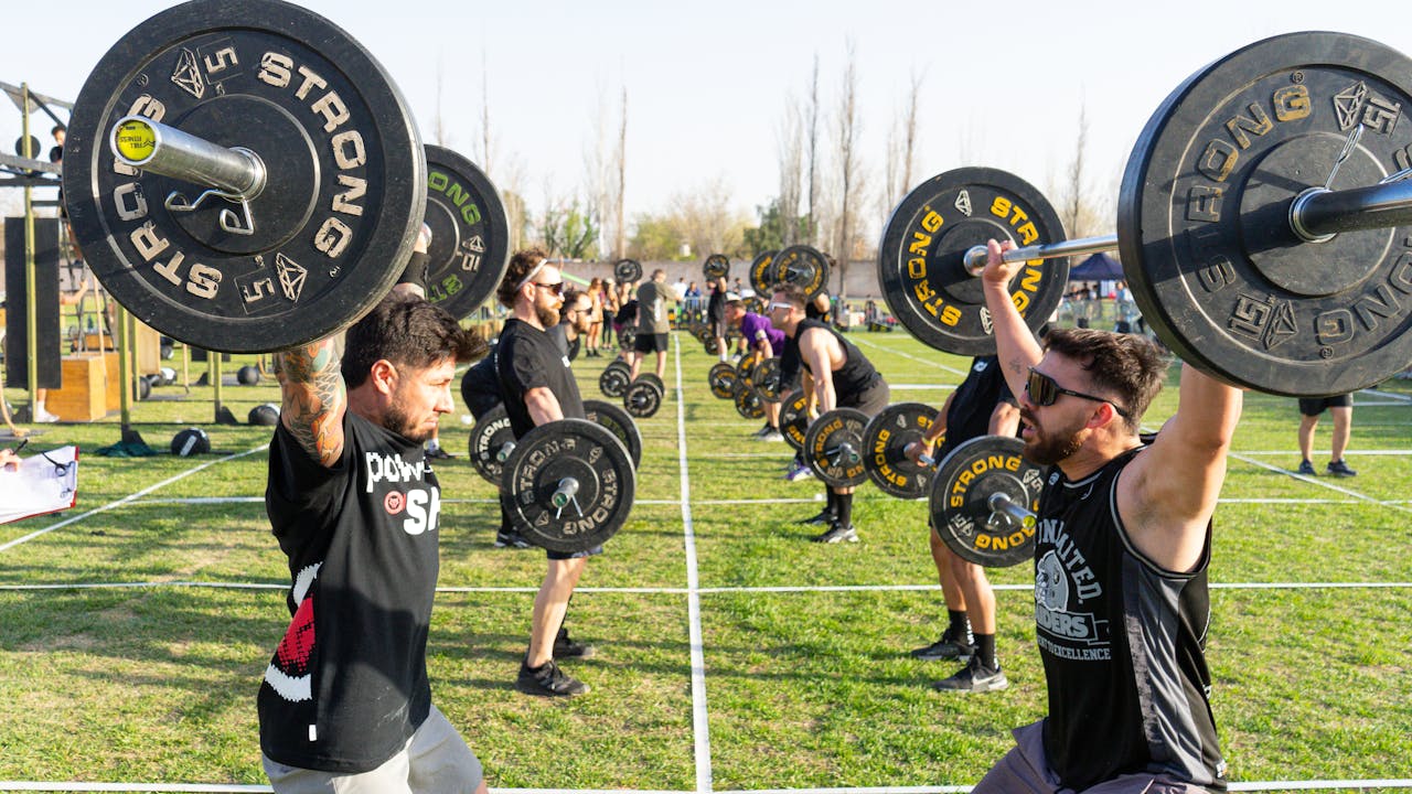 services-01 Strong athletes competing in an outdoor weightlifting event, lifting barbells in daylight.