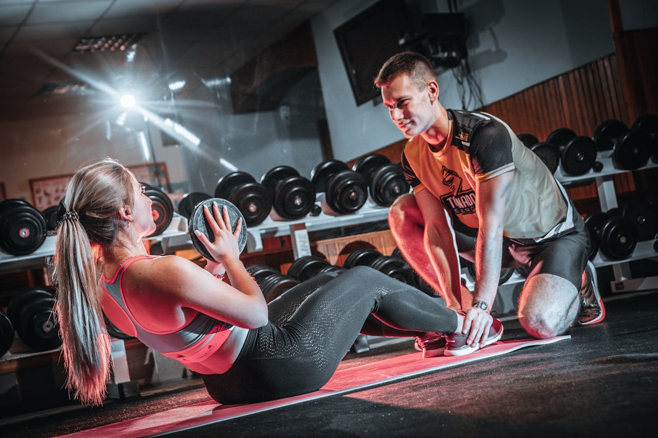 services-03 A personal trainer assists a woman doing sit-ups with a medicine ball in a gym setting.